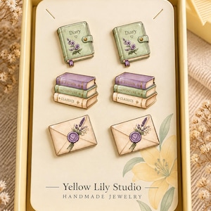 Può includere: Un set di sei orecchini a lobo su un cartoncino con la scritta Yellow Lily Studio Handmade Jewelry. Gli orecchini presentano illustrazioni di diari verdi con lavanda, pile di libri ed buste con sigilli in ceralacca.