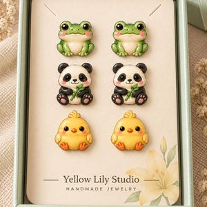May include: Three pairs of stud earrings displayed on a cream-colored card inside a light green box. The earrings feature miniature figures of green frogs, black and white pandas holding bamboo, and yellow chicks. The card reads Yellow Lily Studio.