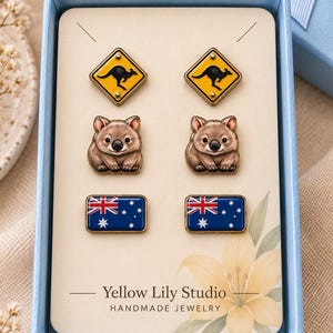 Op de afbeelding: Een set van drie paar oorknopjes op een kaart met de tekst Yellow Lily Studio Handmade Jewelry. De ontwerpen bevatten gele ruitvormige kangoeroe-verkeersborden, bruine wombat-illustraties en rechthoekige Australische vlaggen.