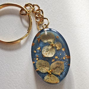 May include: Oval resin pendant keychain with a gold-coloured chain and ring. The pendant features dried flowers and gold flakes set in a deep blue resin. The flowers have a delicate, lacy appearance. A decorative accessory.