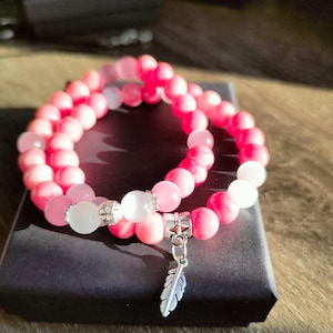 Op de afbeelding: Twee armbanden met roze en witte stenen kralen. De armbanden liggen op een zwarte doos. Eén armband heeft een zilveren veerketting. De kralen zijn een mix van roze en wit, met zilveren accenten.