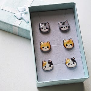 Peut inclure: Un ensemble de trois paires de boucles d'oreilles en forme de chat présentées dans une boîte cadeau bleu clair. Les boucles d'oreilles présentent des illustrations d'un chat gris tigré, d'un roux tigré et d'un tricolore, avec une finition brillante.