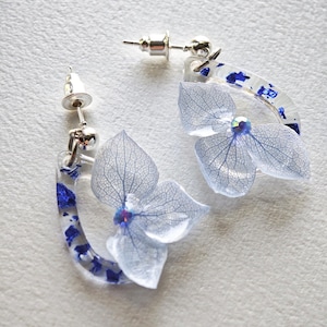 May include: A pair of delicate, handmade earrings featuring light blue hydrangea flowers encased in clear resin. The earrings have a silver post and a curved, clear resin frame with blue accents. The flowers have intricate vein details.