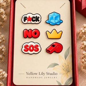 May include: A set of six enamel pins on a display card labeled Yellow Lily Studio. The pins feature a black and white cloud with the text F*CK, a blue ice cube, a red bubble with the text NO, a yellow crown, a red cloud with the text SOS, and a red whistle.