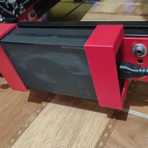 May include: A black power supply unit with a red plastic enclosure is mounted to a black metal frame. A black power cord is plugged into the unit.