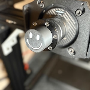 May include: Close-up of a black and silver mechanical device with a carbon fiber pattern. A cylindrical component features a white smiley face. The device is secured with multiple screws and bolts, suggesting a robust construction.