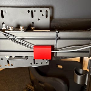 Könnte beinhalten: Ein schwarzer Metall-Schreibtischhalter mit einer roten Kabelmanagementklemme. Der Halter hat eine Reihe von Löchern und einen schwarzen Kabelbinder.