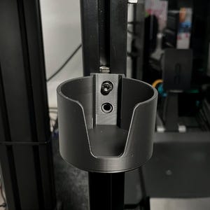 Puede incluir: Un portavasos de plástico negro montado en un poste de metal negro. El portavasos tiene dos agujeros para tornillos en el lateral.