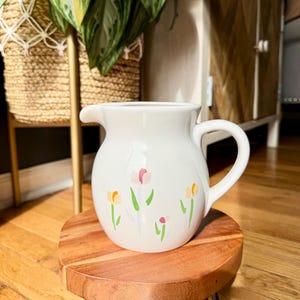 May include: A white ceramic pitcher with a handle and a spout, decorated with painted tulips in pink, yellow, and green. The pitcher sits on a small, round wooden stand with metal legs. The pitcher is a decorative item.