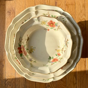 May include: A stack of three octagonal cream-colored ceramic plates with a floral design. The plates have a scalloped edge and a gold trim. The floral pattern includes red, yellow, and purple flowers with green leaves.