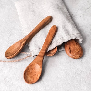 May include: Several wooden spoons with a natural finish are arranged on a light-colored surface. The spoons have a smooth, curved design and are made of a light brown wood. A linen cloth is partially visible in the background, adding a rustic touch to the composition.