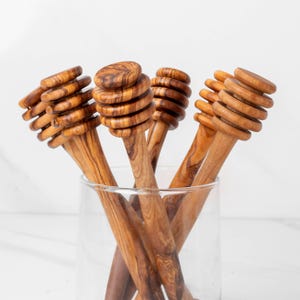 May include: A collection of wooden honey dippers displayed in a clear glass. The dippers are made of light brown wood with a spiral design on the handle and a rounded top. The wood grain is visible, adding to the natural aesthetic.
