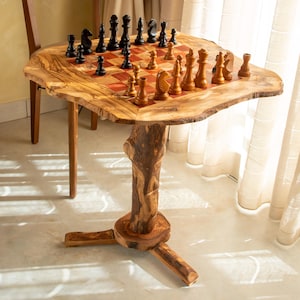 May include: A live edge wooden pedestal table featuring a built-in chessboard top with alternating light and dark squares. The table is supported by a thick, textured tree trunk base with three feet. A full set of black and light brown chess pieces.