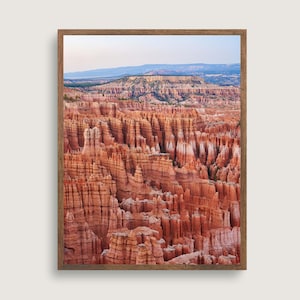 Puede incluir: Una vista panorámica del Parque Nacional Bryce Canyon en Utah, que presenta una vasta extensión de hoodoos de roca roja, una formación geológica creada por la erosión. Los hoodoos son altos, delgados y en forma de aguja, con una variedad de colores y texturas. El cielo es azul claro y el sol brilla.