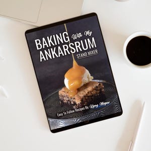 May include: A tablet displays a cookbook cover titled "Baking With My Ankarsrum Stand Mixer." The cover features a brownie topped with ice cream and caramel sauce. A cup of coffee and a pen are also visible.
