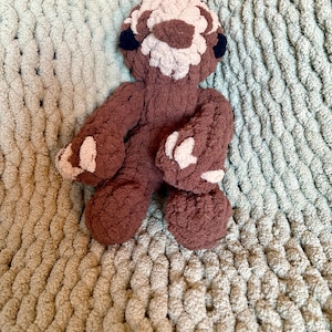 May include: A brown and pink crocheted sloth plush toy with black eyes. The toy is sitting on a light green knitted blanket.