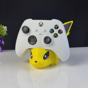 Puede incluir: Un mando blanco de Xbox descansa sobre una figura amarilla de dibujos animados. El mando tiene joysticks negros y botones de colores. La figura tiene ojos negros y una pequeña nariz negra. El fondo es blanco y negro.