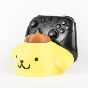May include: A yellow character-shaped controller stand with a brown top accent, featuring a simple black face design with two eyes and a mouth. A black Nintendo Switch Pro controller is positioned securely within the stand's rear slot.