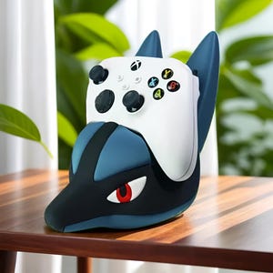 Op de afbeelding: Een witte Xbox-controller rust in een blauw-zwarte Lucario-thema controllerstandaard. De standaard heeft een gestileerde Lucario-kop met een rood oog en blauwe accenten. De controller heeft zwarte knoppen en gekleurde actieknoppen.