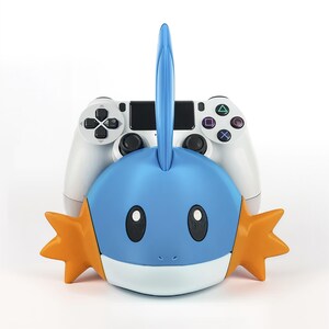 May include: A blue and orange Mudkip figure with a white base, positioned in front of a white PlayStation 4 controller. The Mudkip has black oval eyes and orange star-shaped fins. The controller has black buttons and directional pad.