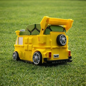 May include: A yellow toy van with a green top and a large spoiler. The van has gray wheels and a spare tire on the back. The back of the van has the letters "TM-NT" on it. The toy is sitting on green grass.