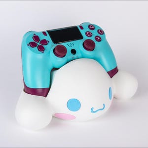 May include: A turquoise and dark purple video game controller resting on a white, rounded character-shaped stand. The controller features a central black touchpad, two analog sticks, and standard action buttons. The stand has blue eyes and pink cheeks.