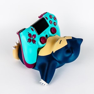May include: A teal and purple PlayStation 4 controller rests on a blue, tan, and white Snorlax figure. The controller has a black screen and purple buttons. The Snorlax figure is lying down.