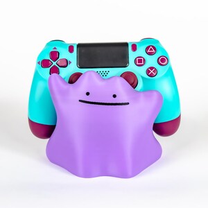 May include: A purple Ditto figure in front of a teal and maroon game controller. The controller has black accents and colored buttons. The Ditto figure has a simple smiling face.