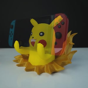 May include: A yellow Pikachu figure with outstretched arms, emerging from a stylized flame base. A red and black Nintendo Switch controller is visible behind the figure. The background is dark.