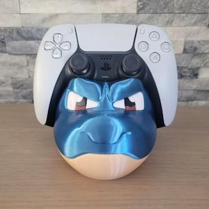 May include: A blue and tan 3D-printed controller holder shaped like a cartoon character's head, with a white and black game controller resting on top. The holder has a glossy finish and is designed to hold a gaming controller.