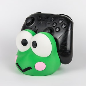 May include: A green frog-shaped controller stand featuring large white eyes with black pupils, pink circular cheeks, and a black V-shaped mouth. A black Nintendo Switch Pro controller is resting securely inside the holder, displaying its buttons.