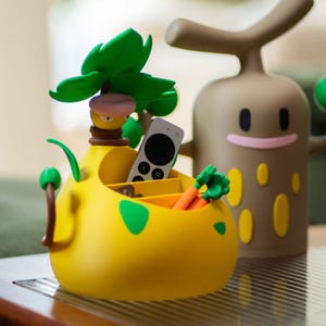 Può includere: Un organizer giallo in stile cartone animato a forma di albero con accenti verdi e una cima verde e frondosa. Contiene un telecomando e piccole carote arancioni. Una figura di cartone animato marrone è dietro.
