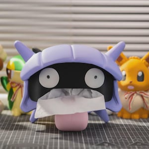 Op de afbeelding: Een verzameling kleurrijke Pokémon-karakterbeeldjes, waaronder een tissuebox in de vorm van een paarse Shellder. De box heeft een zwart gezicht met witte ogen en een witte tissue die eruit steekt. Andere beeldjes zijn Eevee en een bruin konijn.