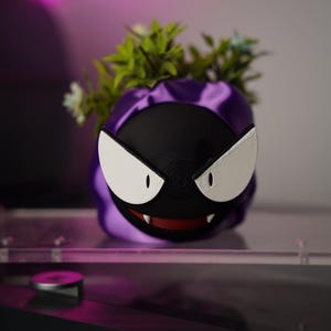 Puede incluir: Una maceta impresa en 3D con la forma de un personaje de Pokémon Gastly. La maceta presenta una cara negra con ojos blancos y una boca roja, rodeada de tela morada. Hay plantas verdes y pequeñas flores blancas en el interior.