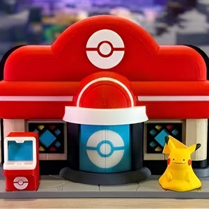 May include: A red and white Pokemon Center building playset featuring a central entrance with a circular logo, two side windows with blue diamond patterns, a small red kiosk, and a yellow Pikachu figure standing on a gray base platform.