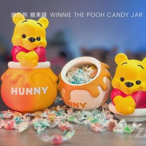 May include: A Winnie the Pooh candy jar featuring a yellow bear with a red shirt emerging from a round orange pot labelled HUNNY. The set includes a matching open jar filled with colourful wrapped sweets and a separate lid with the bear figure.