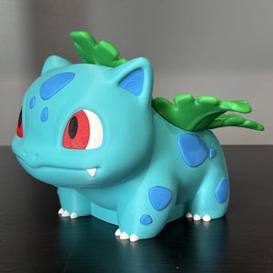 May include: A light blue creature figurine with dark blue spots, red eyes, and a green plant bulb structure on its back. The figure has four legs with small white claws and a small white tooth visible in its mouth, rendered in a smooth finish.