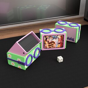 May include: Three colorful boxes with a unique design, featuring a green base with white daisy accents, pink tops, and purple accents. The boxes have a glasses-like design on the sides. One box is open, revealing a black interior. A white die is on the black surface.