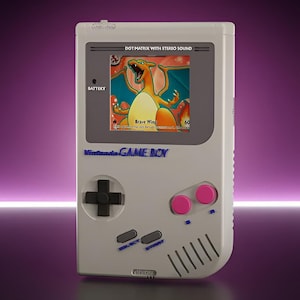 Peut inclure: Une console de jeu vidéo portable gris clair avec un écran affichant un personnage Dracaufeu. L'appareil comprend une croix directionnelle noire, deux boutons d'action roses et des boutons Select et Start gris. Le texte Nintendo GAME BOY est imprimé en bleu, avec les mentions DOT MATRIX WITH STEREO SOUND et BATTERY visibles.