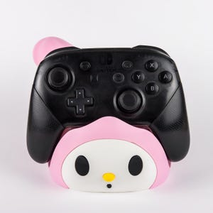 May include: A black Nintendo Switch Pro controller resting in a pink and white character-themed charging dock. The controller features standard black buttons, joysticks, and the Nintendo Switch logo. The dock has a rounded pink hood and white face.