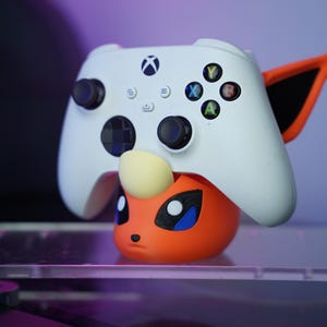 May include: A white Xbox controller rests on an orange and yellow character-themed stand. The controller has black joysticks and colored buttons. The stand has black, blue, and white details. The background is blurred with purple and pink hues.