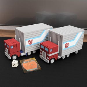 May include: Two red and white toy semi-trucks with grey cargo trailers featuring blue stripes and red Autobot logos. A single white six-sided die and a rectangular Magic The Gathering trading card with an orange background are placed in front.