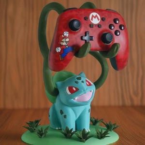 Könnte beinhalten: Ein roter Videospiel-Controller mit Mario-Grafik ruht auf einer grünen, rankenartigen Struktur, die von einer blauen Bulbasaur-Figur getragen wird. Die Basis ist ein grüner Kreis mit kleinen Pflanzendetails. Der Controller hat schwarze Knöpfe und ein weißes 'M'-Logo.
