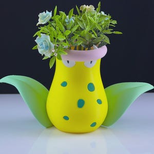 Puede incluir: Una maceta amarilla caprichosa con un diseño de dibujos animados, con lunares verdes y grandes hojas laterales verdes. La maceta contiene flores artificiales con rosas azules y blancas y follaje verde. Un artículo decorativo único.