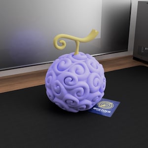 Könnte beinhalten: Eine hellviolette, kugelförmige Frucht mit einem gelben Stiel und einer Karte mit der Aufschrift "ONE PIECE". Die Frucht hat eine strukturierte Oberfläche mit wirbelnden Mustern. Die Karte ist blau mit goldenen Akzenten.