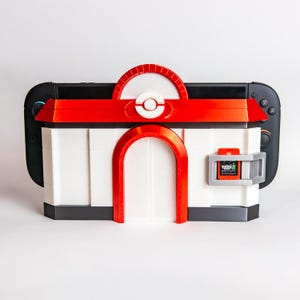 May include: A white and red 3D-printed case for a Nintendo Switch, designed to resemble a building from the Pokémon series. The case has a red roof, archway, and accents, with a game cartridge slot. The Nintendo Switch is black.