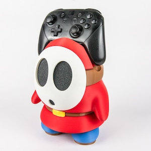 Op de afbeelding: Een rood, wit en blauwe controllerstandaard in de vorm van een personage die een zwarte Nintendo Switch Pro-controller vasthoudt. De standaard heeft een wit ovaal masker met twee zwarte oogopeningen, een rood lichaam, een bruine riem en blauwe voeten.