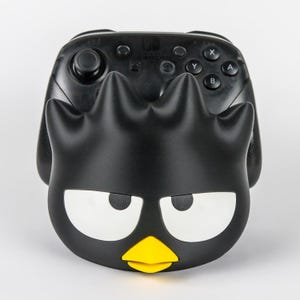 May include: A black controller stand shaped like a cartoon character with three spiky points on top, large white eyes with black pupils, and a small yellow beak. The stand holds a black wireless game controller with standard buttons and joysticks.