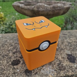 May include: A square orange storage box featuring a graphic of a creature's eyes and mouth on the top lid. The front of the box displays a black horizontal band with a circular white button in the centre, resembling a spherical container design.
