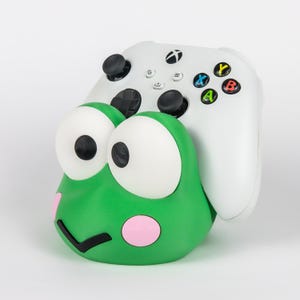 May include: A green frog-shaped controller stand featuring large white eyes, pink circular cheeks, and a black mouth. A white wireless gaming controller with black joysticks and multicolored action buttons is resting securely in the stand.